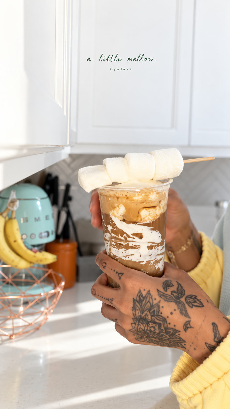 A creamy marshmallow latte held in tattooed hands, topped with three marshmallows in a bright white kitchen